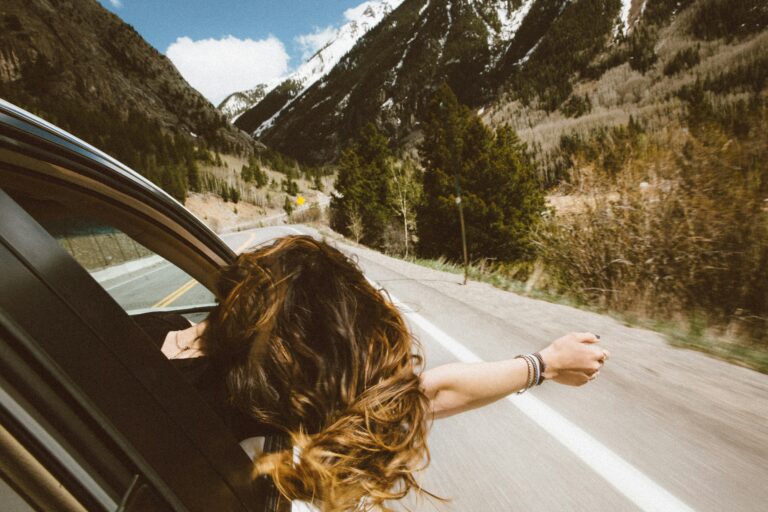 A blonde woman on a road trip seen from the back seat, her head sticking out and her right arm outstretched as she looks on at the winding road ahead,